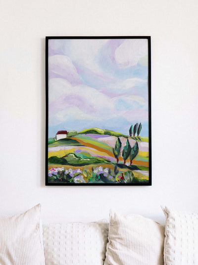 Framed artwork of a colorful landscape with rolling hills and trees on a white wall above a sofa.