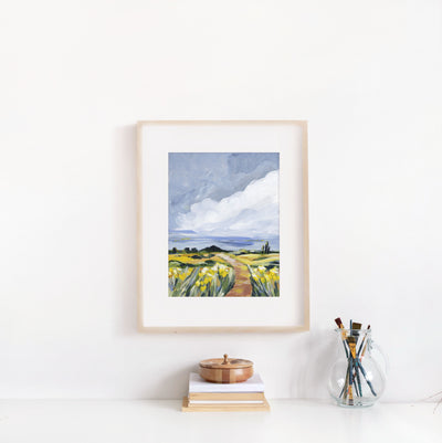 Framed artwork of a landscape with a path and fields on a white wall.