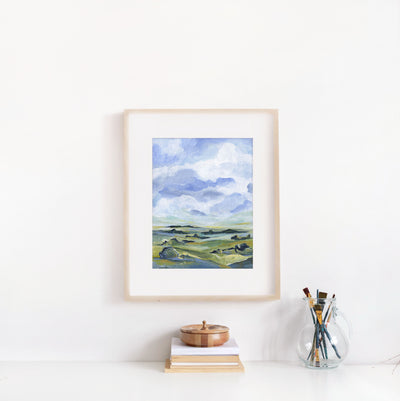 Framed landscape painting on a white wall with books and a glass jar of paintbrushes below.