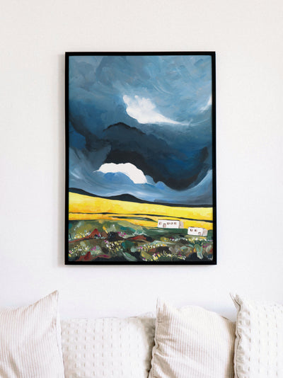 Framed painting of a landscape with dark sky and illuminated buildings on a white wall above a sofa.