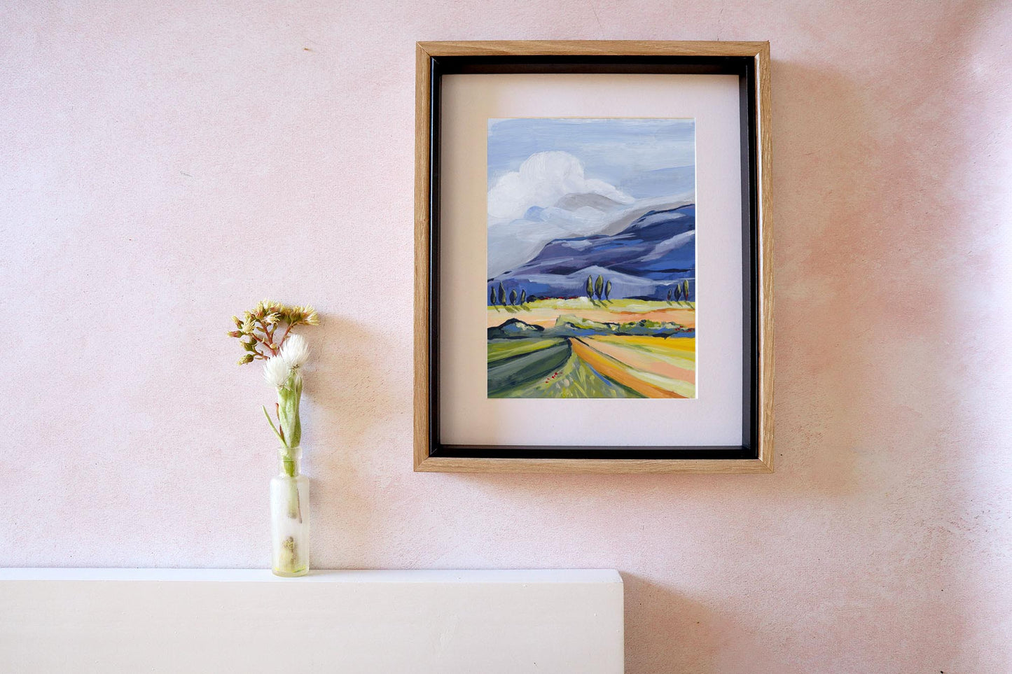 Framed painting of a landscape on a wall with a vase of flowers beside it.