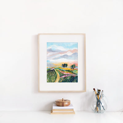 Framed artwork of a landscape with mountains and fields on a white wall.