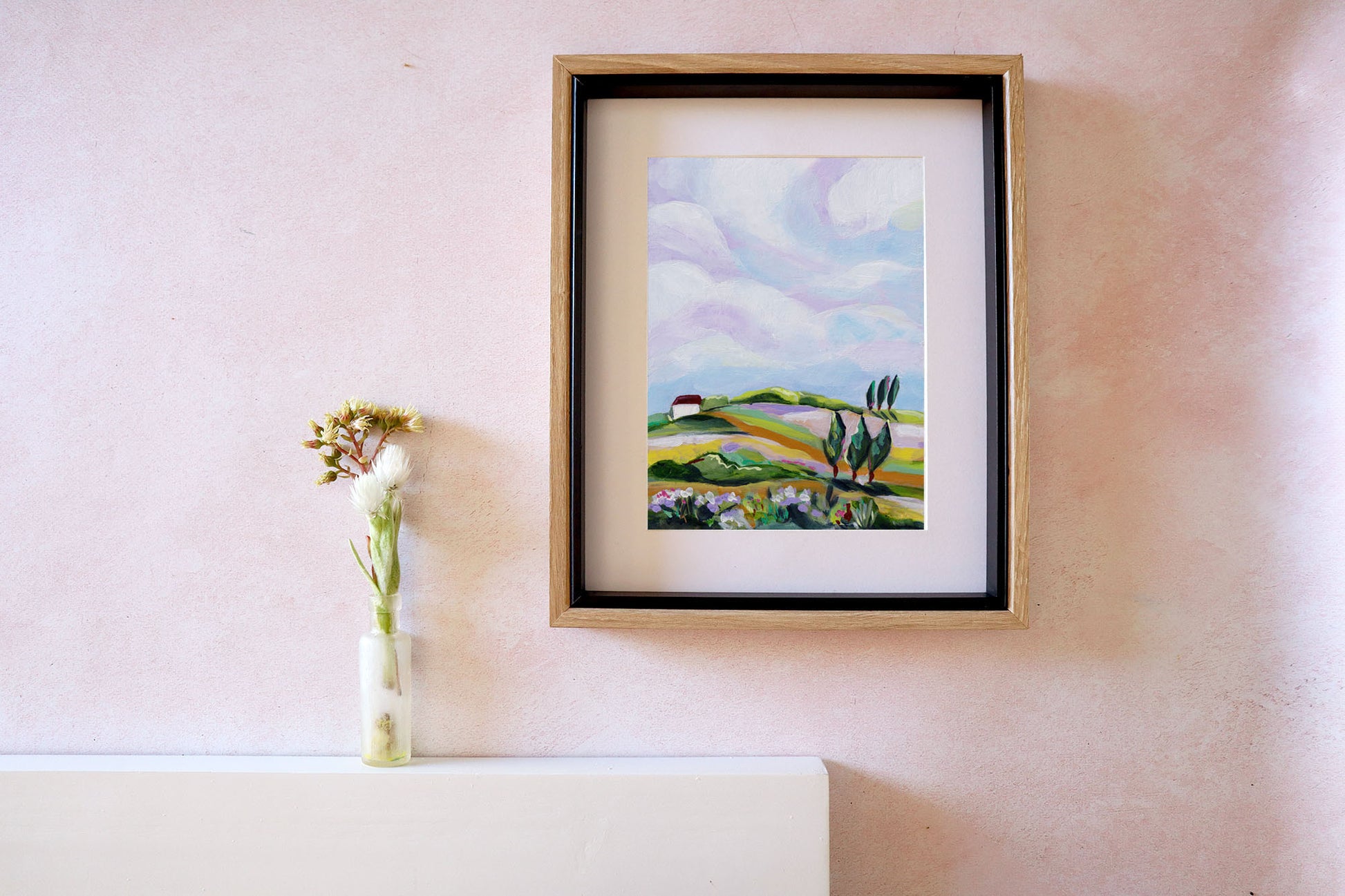 Framed painting of a landscape on a wall next to a vase with flowers.