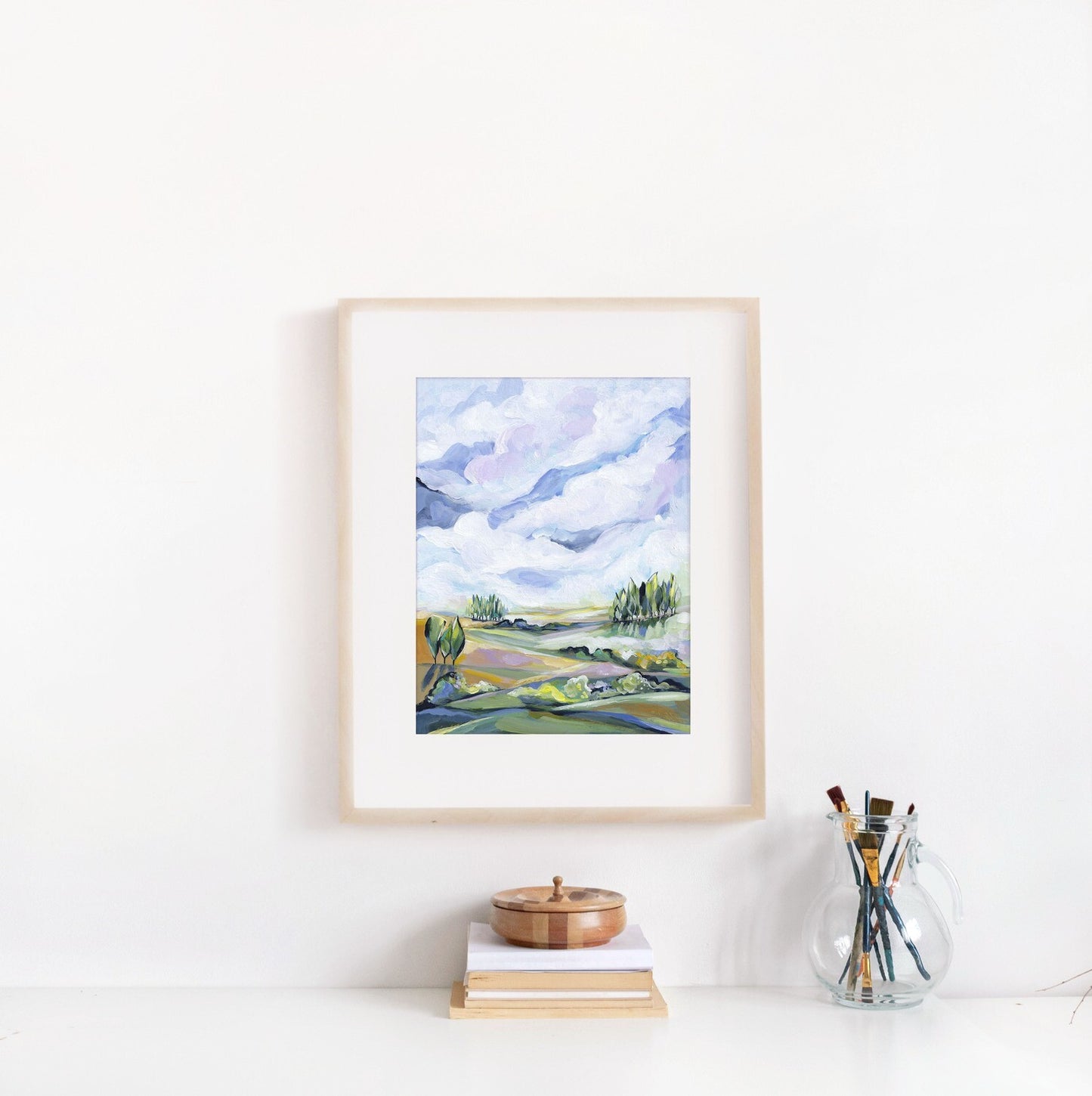 Framed landscape painting on a white wall with books and a glass jar of paintbrushes below.