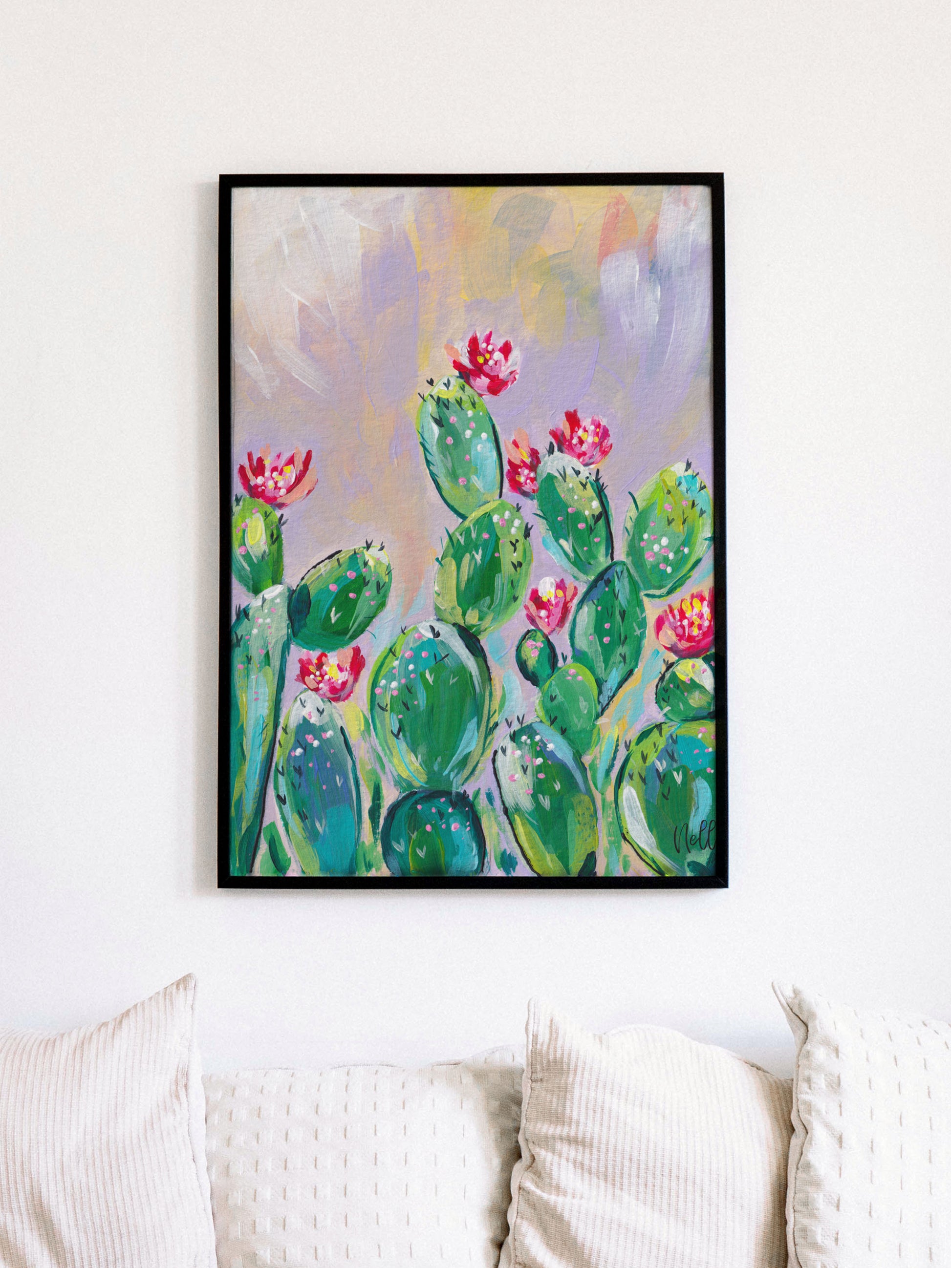 Framed artwork of cacti with pink flowers on a white wall above a sofa.
