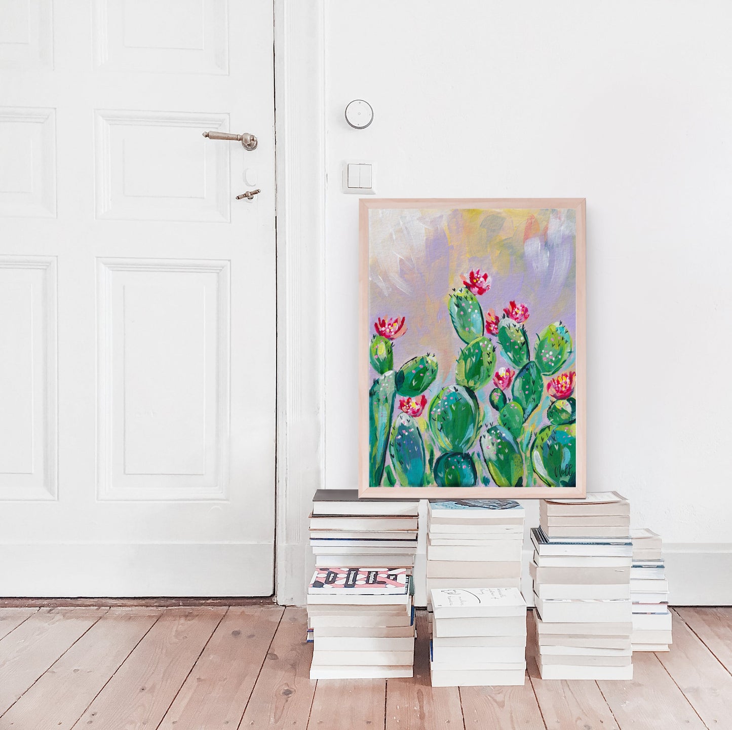Colorful cactus painting leaning against a white wall with stacks of books on a wooden floor.