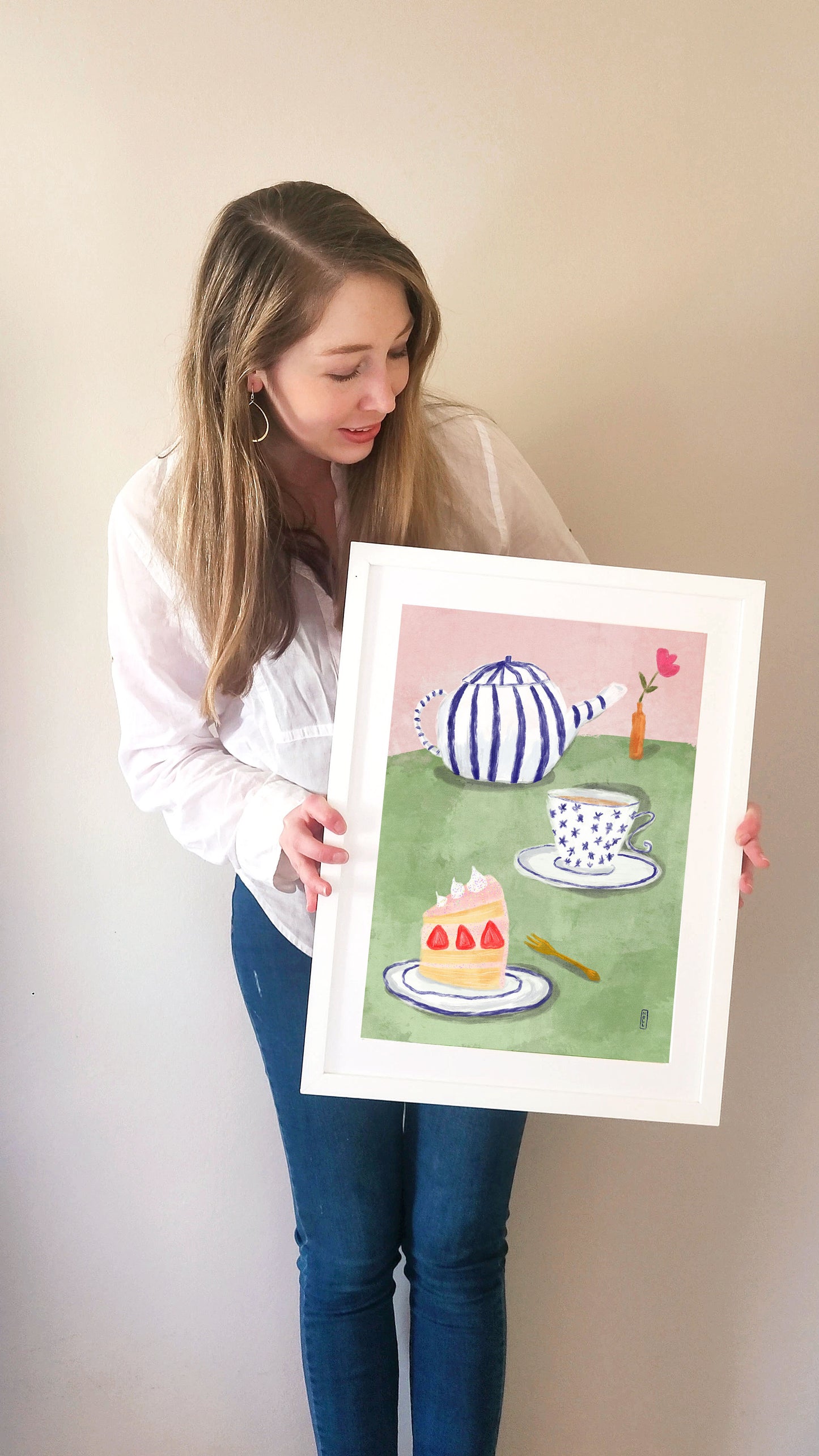 Woman holding a framed illustration of a tea party scene with a teapot, cake, and cups.