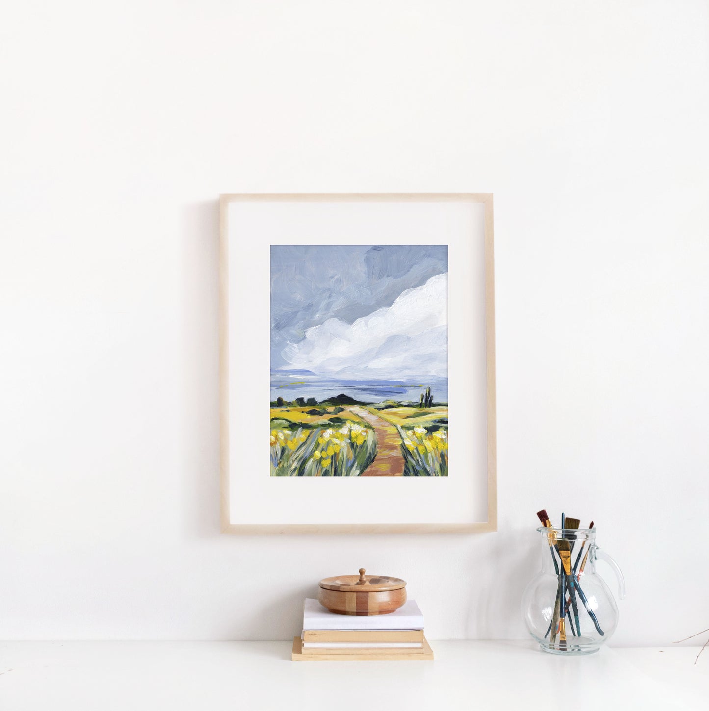 Framed artwork of a landscape with a path and fields on a white wall.