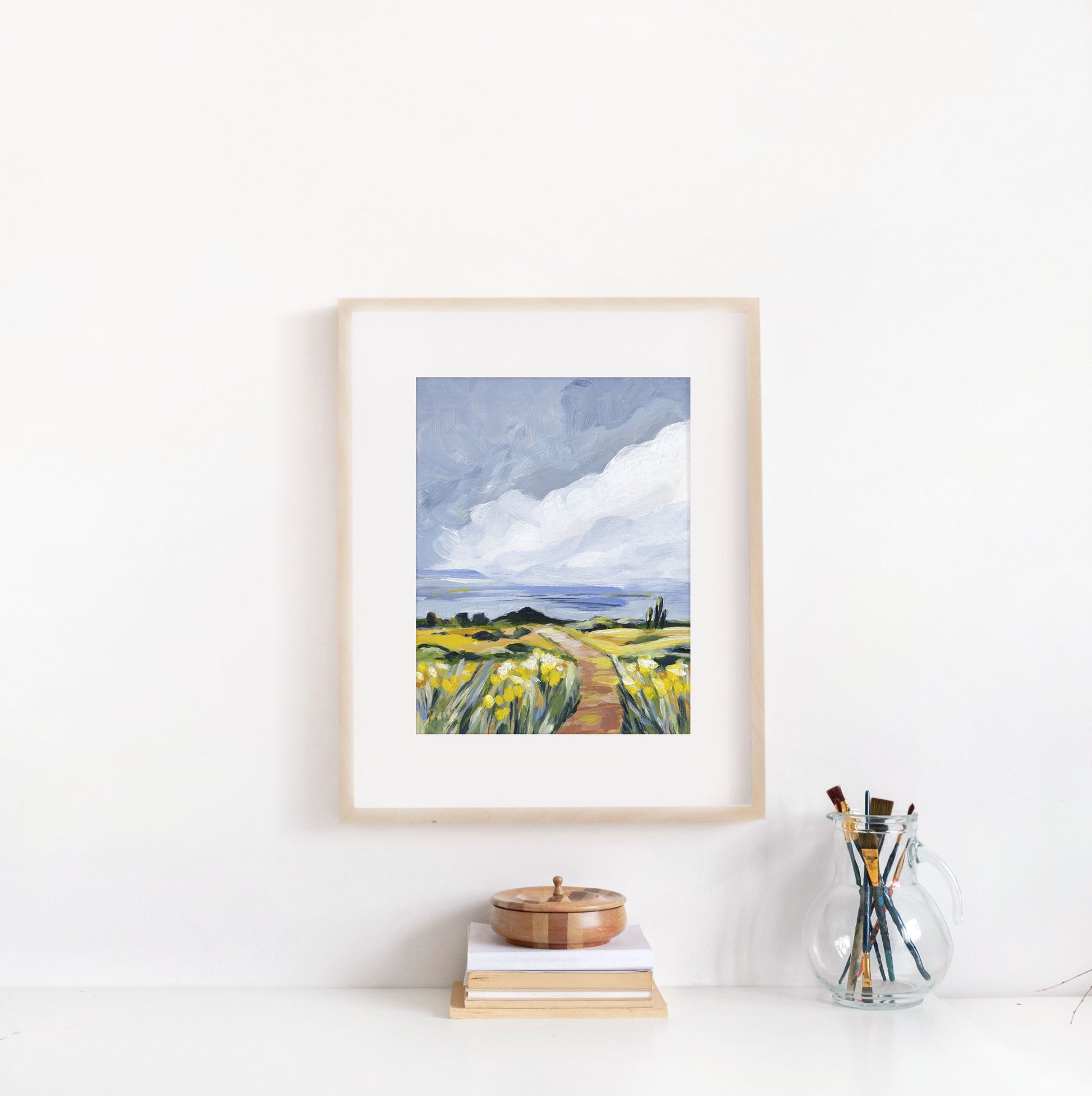 Framed artwork of a landscape with a path and fields on a white wall.