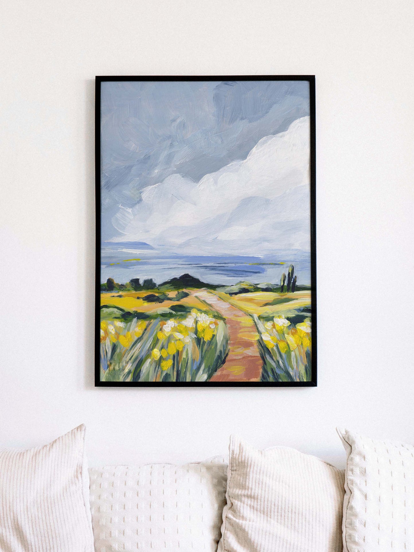Framed artwork of a landscape with a path and yellow flowers on a white wall above a sofa.