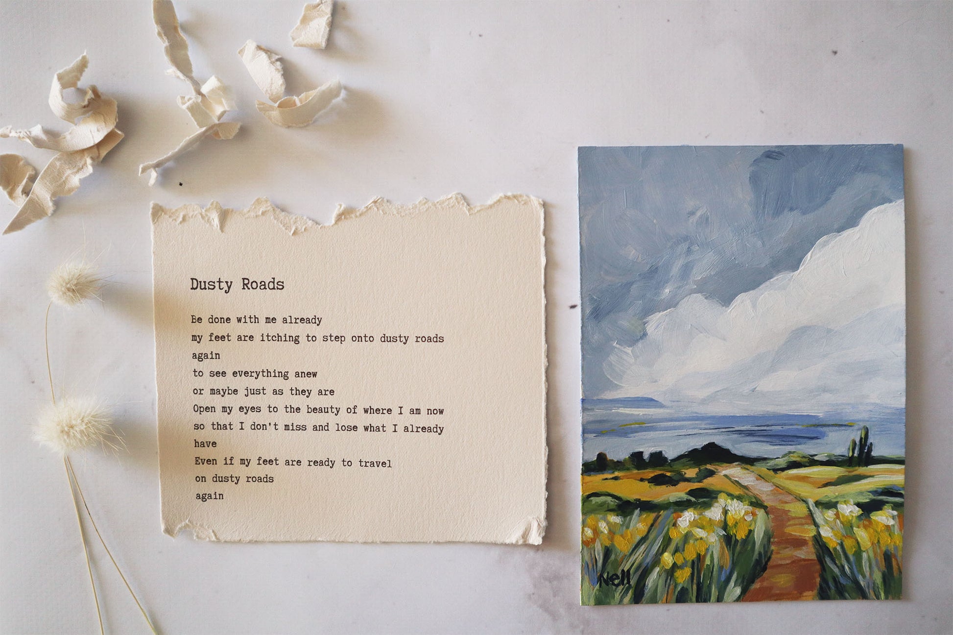 Poem titled 'Dusty Roads' on a piece of paper next to a painting of a landscape with a path and flowers.