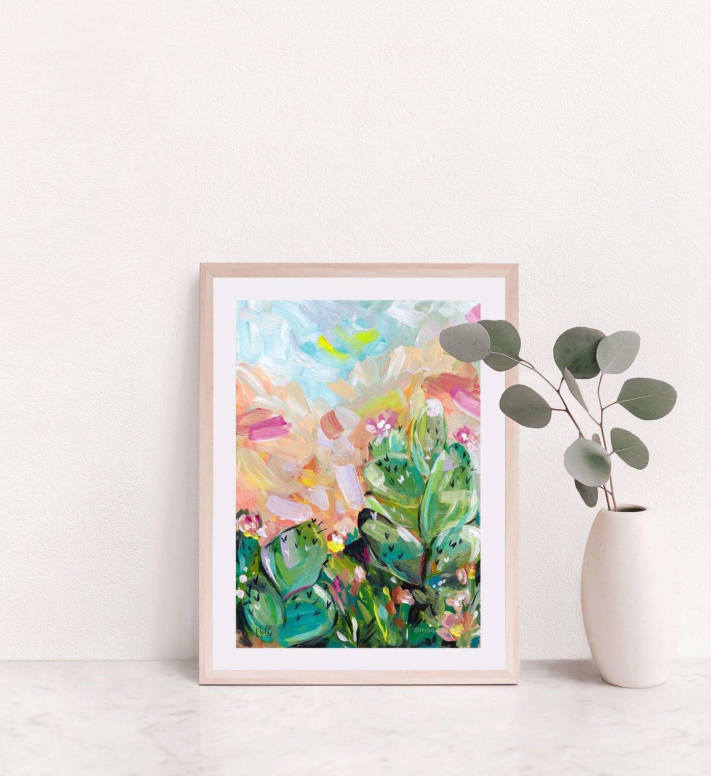 Framed abstract art print on a white wall with a vase of eucalyptus.
