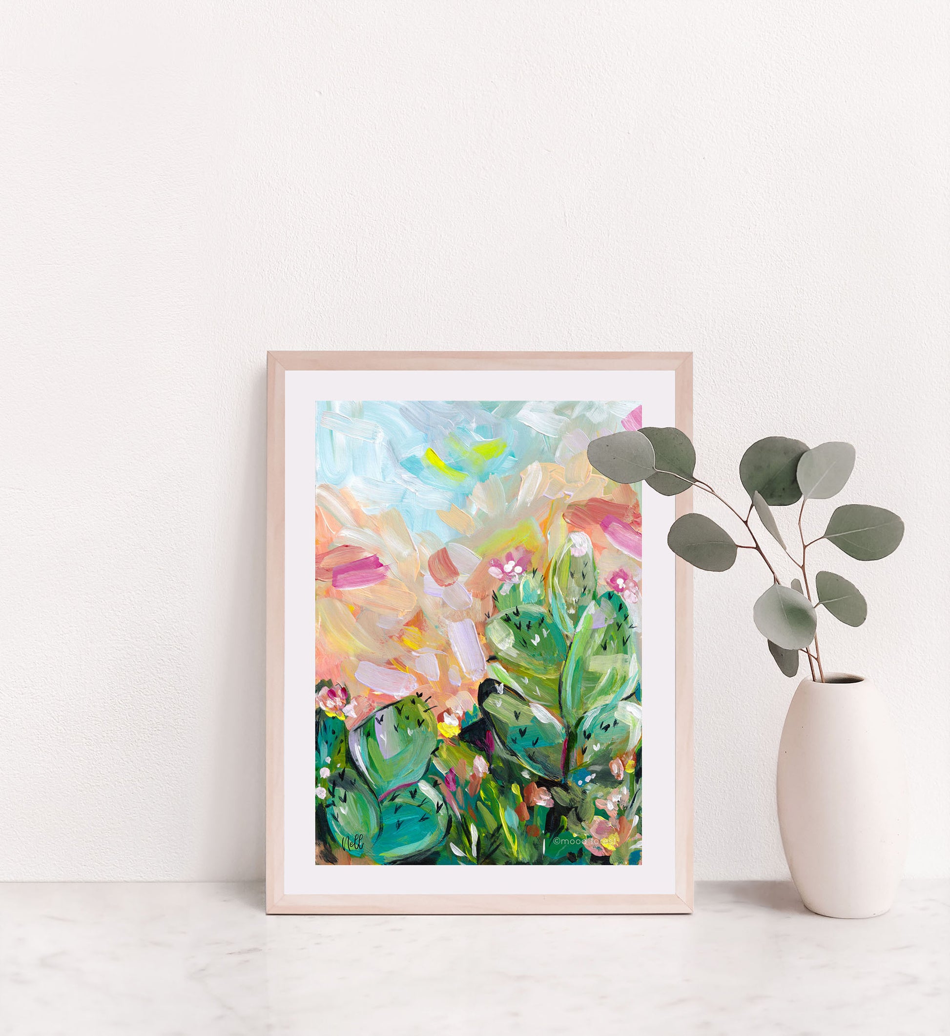 Framed abstract art print on a white wall with a vase of eucalyptus.