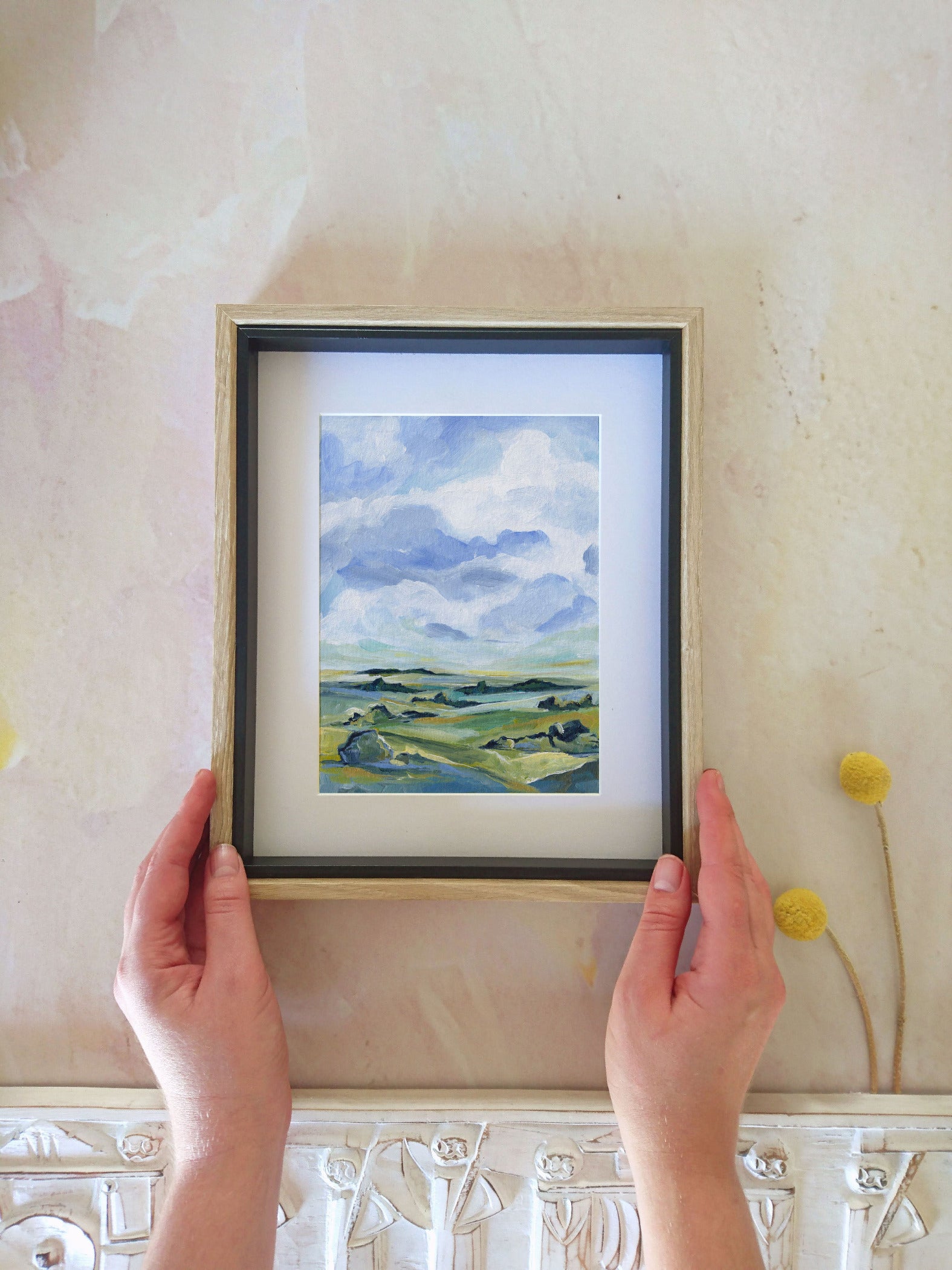 Framed painting of a landscape held by hands against a textured wall.