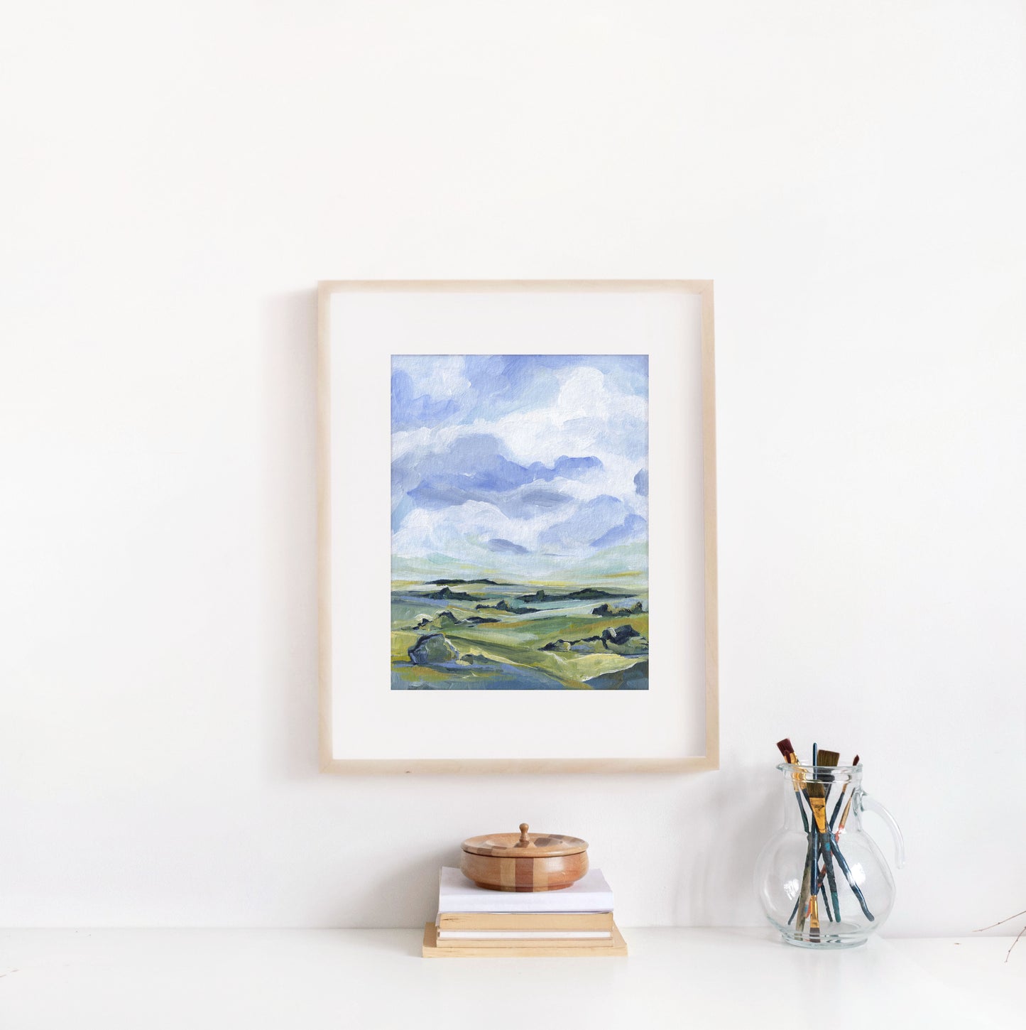 Framed landscape painting on a white wall with books and a glass jar of paintbrushes below.