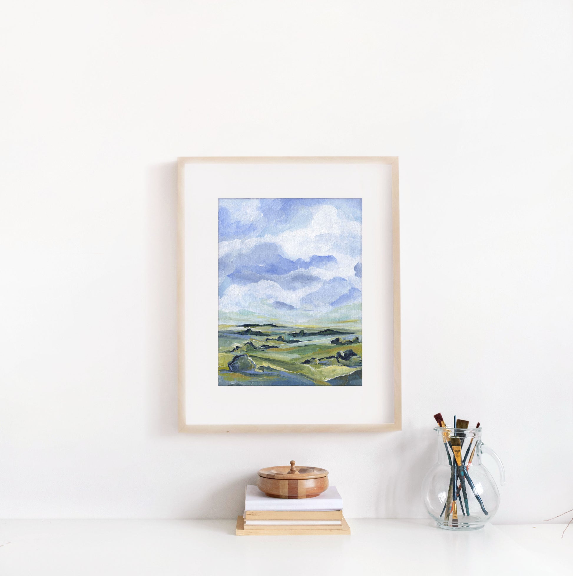 Framed landscape painting on a white wall with books and a glass jar of paintbrushes below.