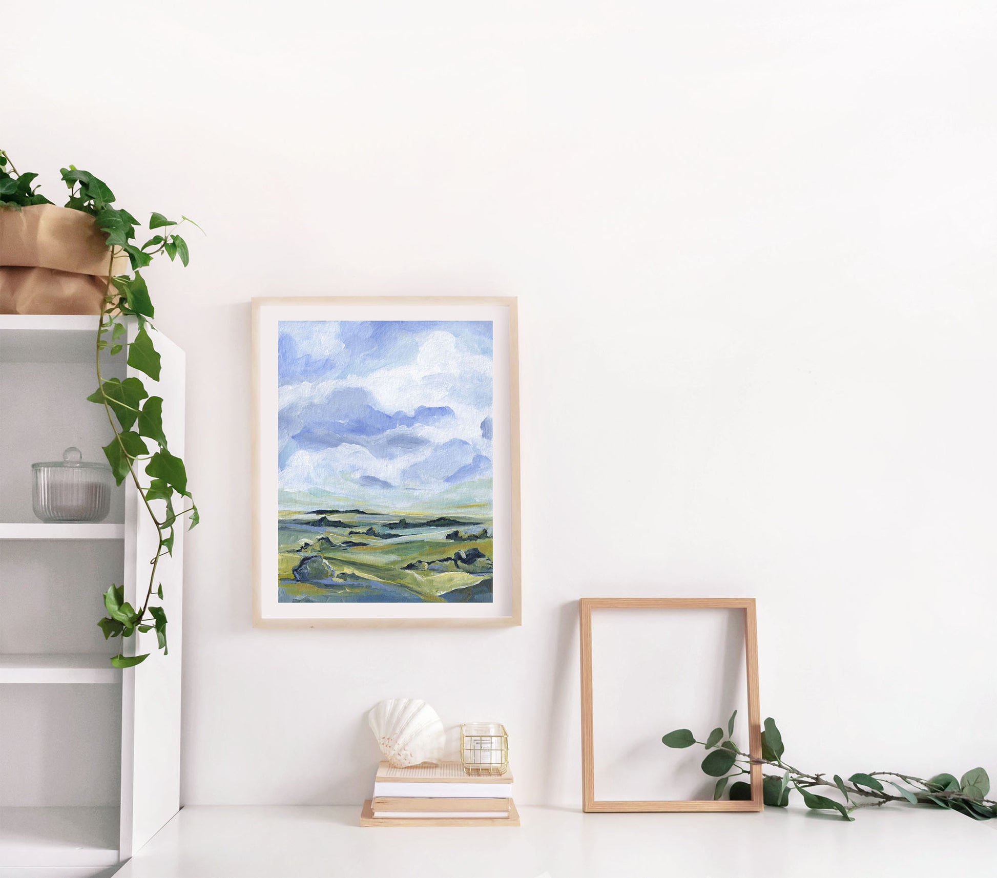 Framed landscape painting on a white wall with a shelf and plant in the background