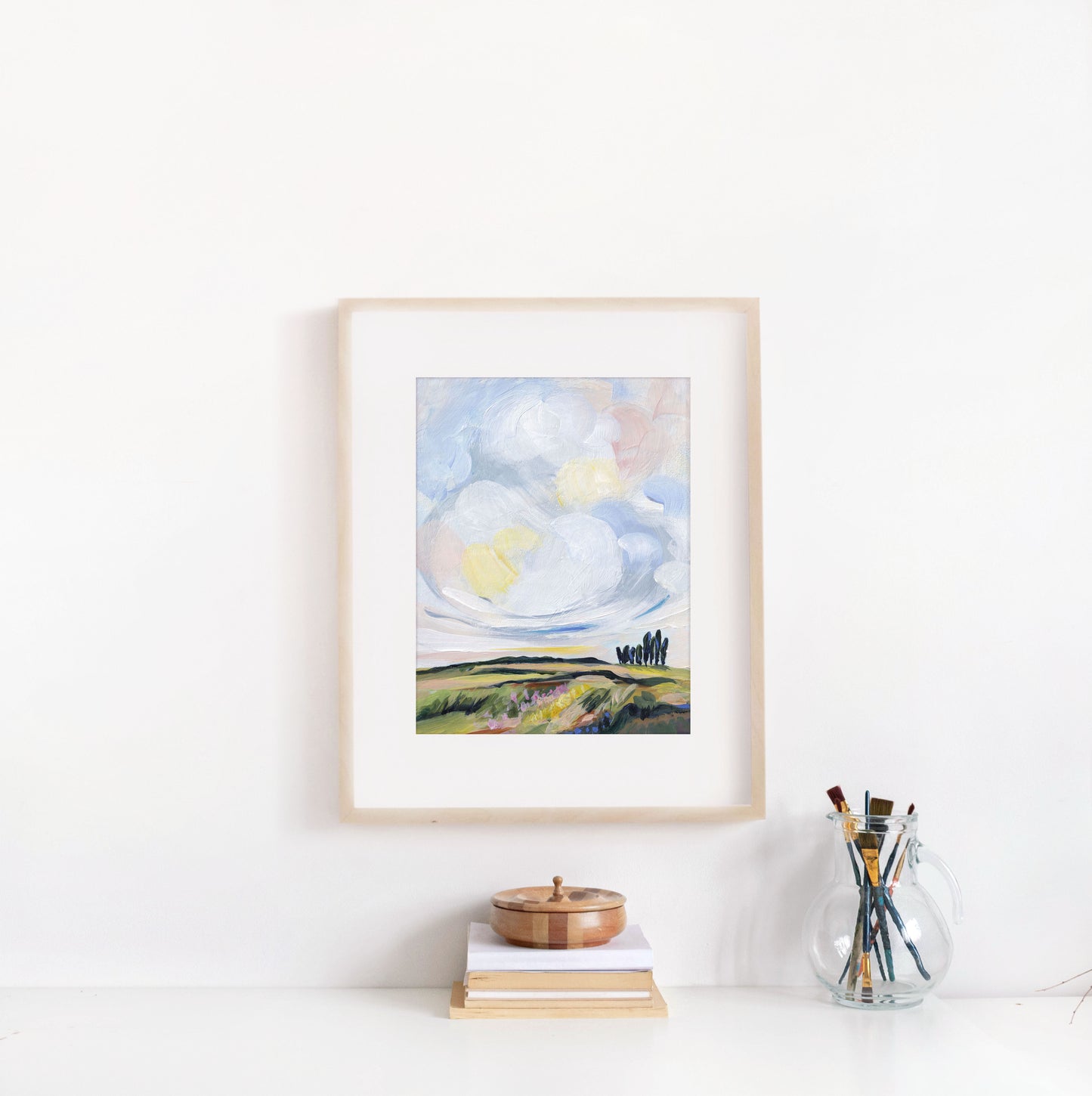 Framed artwork of a landscape with a sun on a white wall, below a shelf with books and a glass jar.