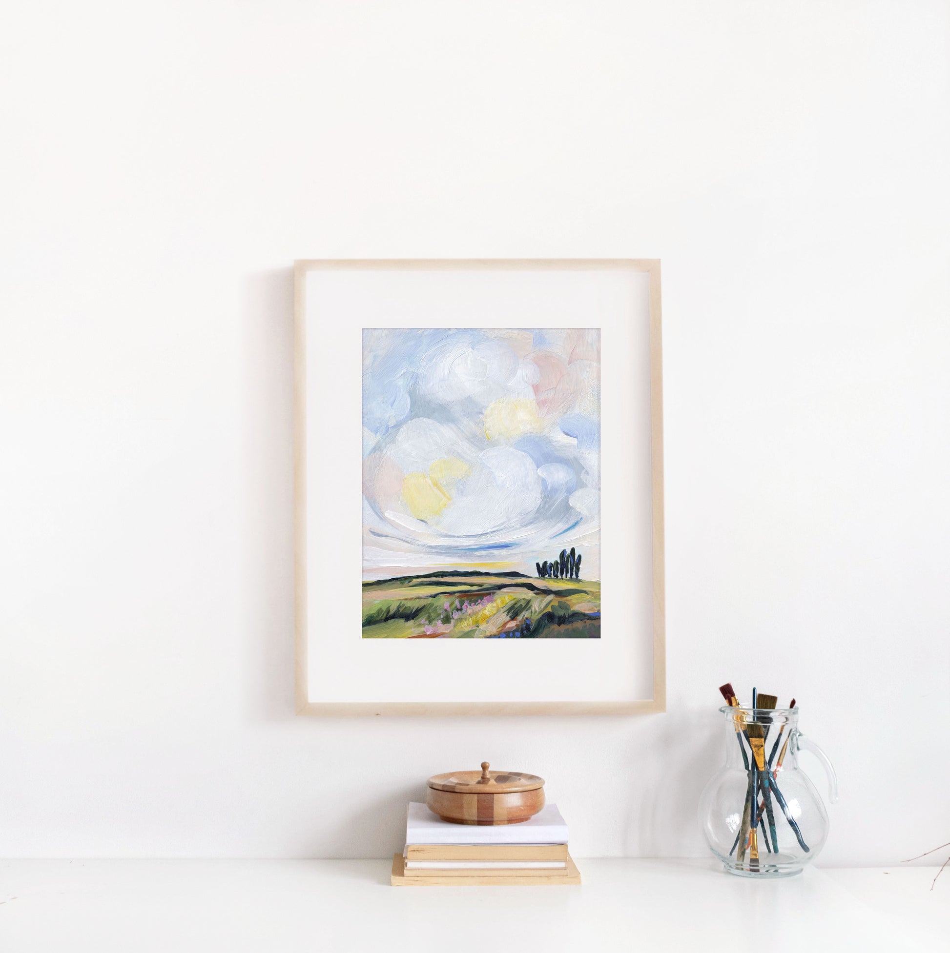 Framed artwork of a landscape with a sun on a white wall, below a shelf with books and a glass jar.