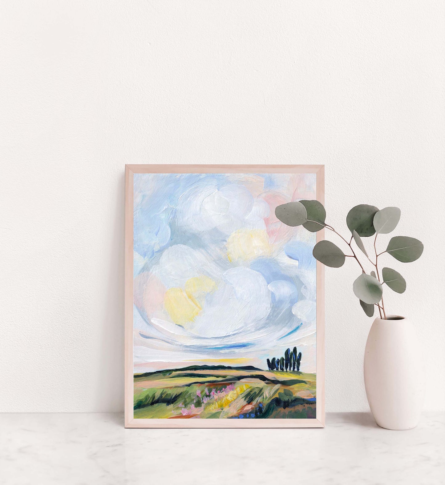 Framed artwork of a landscape with clouds and trees on a white wall.
