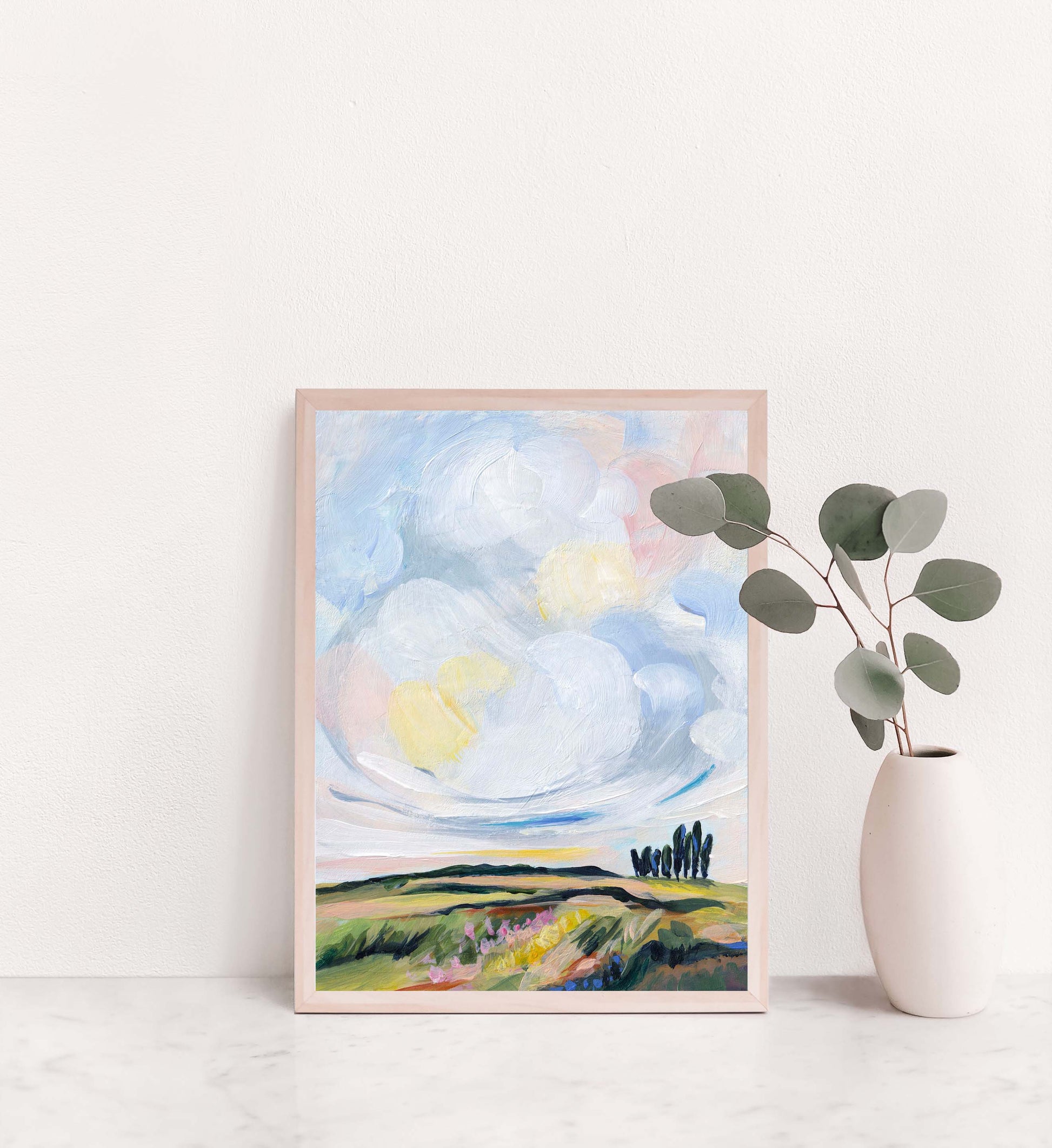 Framed artwork of a landscape with clouds and trees on a white wall.