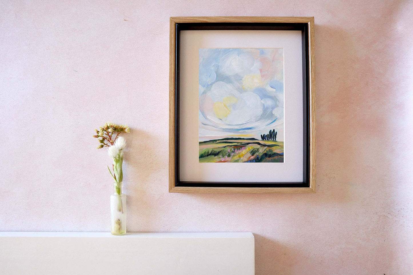 Framed painting of a landscape with a cloud on a wall next to a vase with flowers.