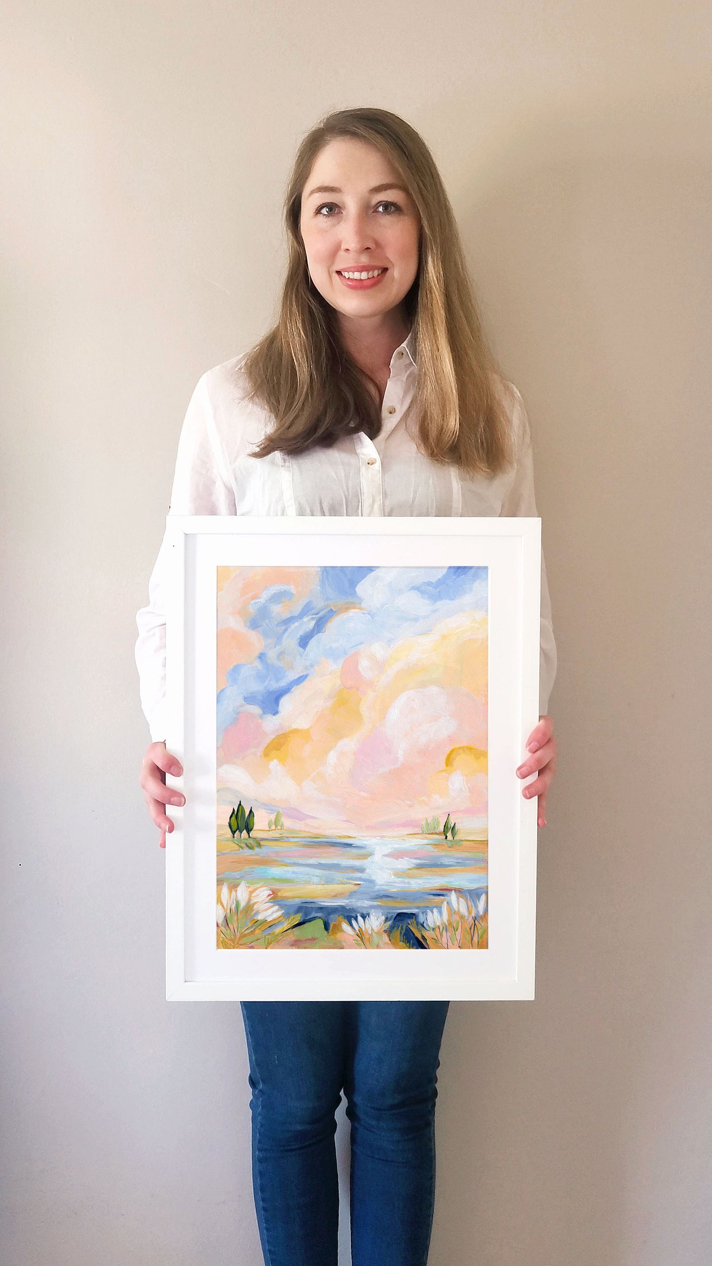 Woman holding a framed watercolor painting of a landscape with a lake and trees.
