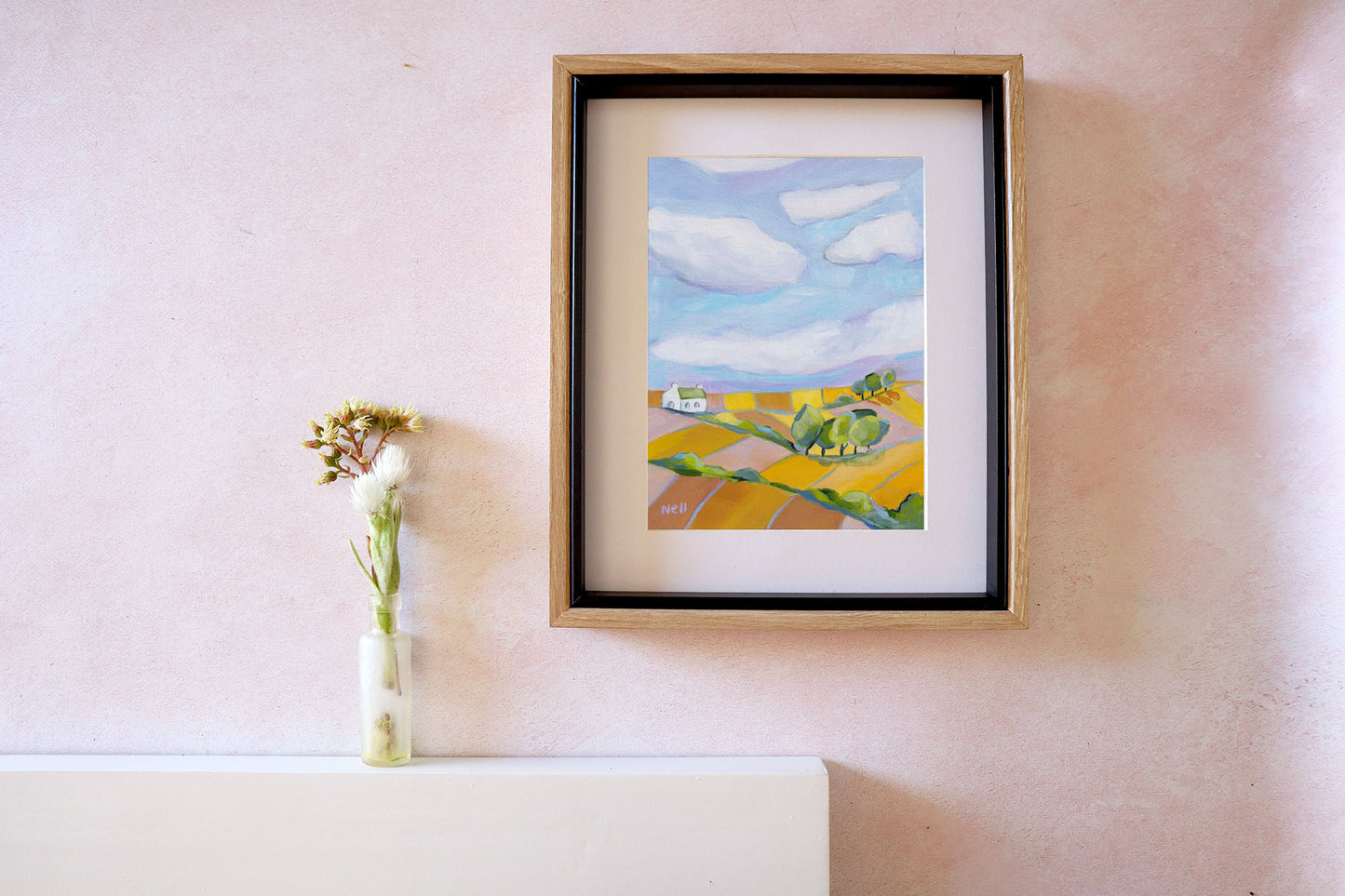 Framed painting of a landscape with fields and a house on a wall next to a vase with flowers.
