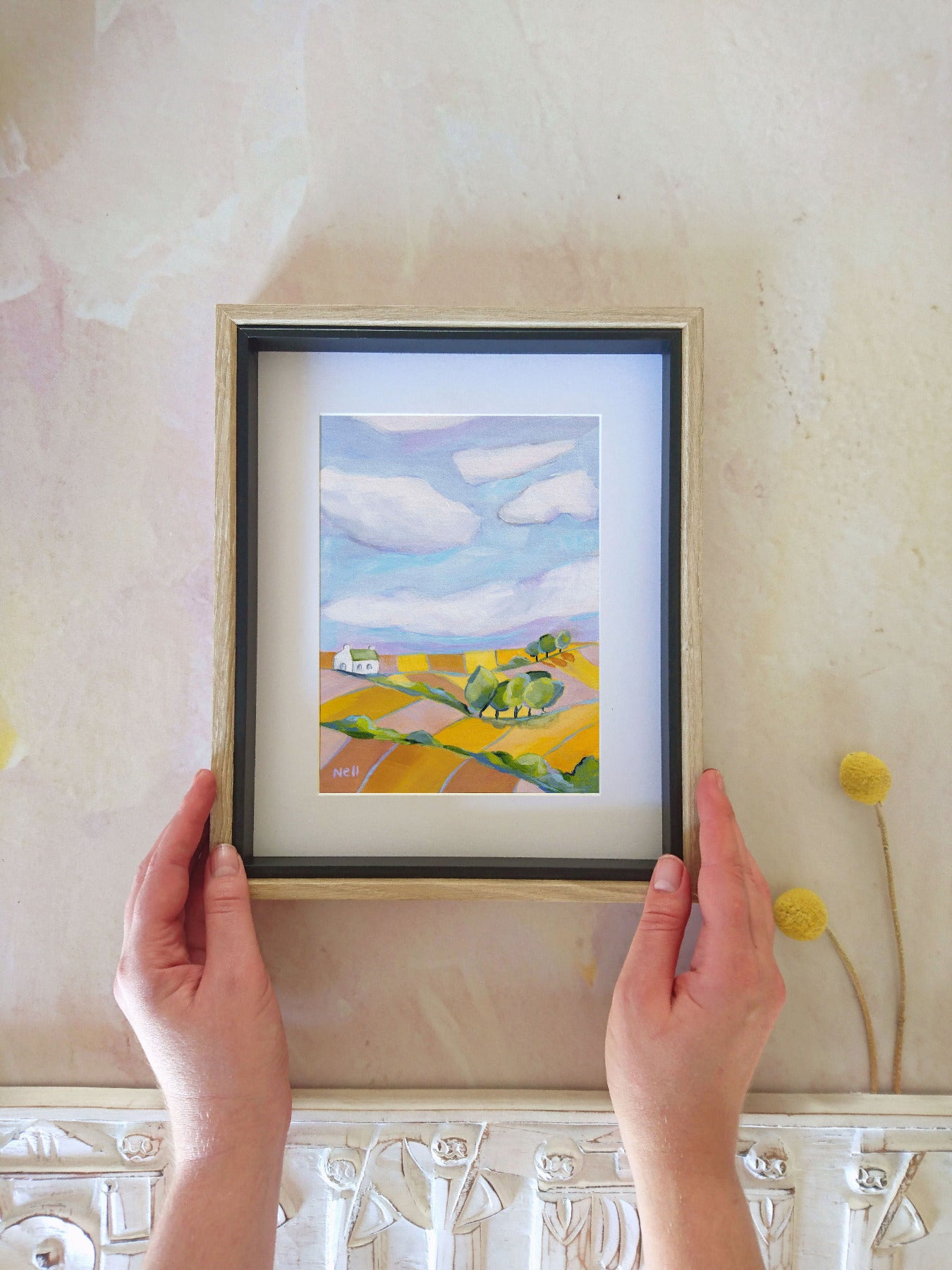Framed painting of a landscape with fields and a house, held by hands against a textured wall.
