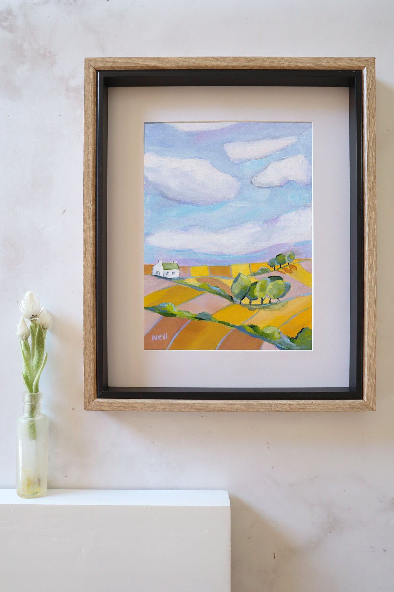 Framed painting of a landscape with a blue sky and yellow field on a white wall.