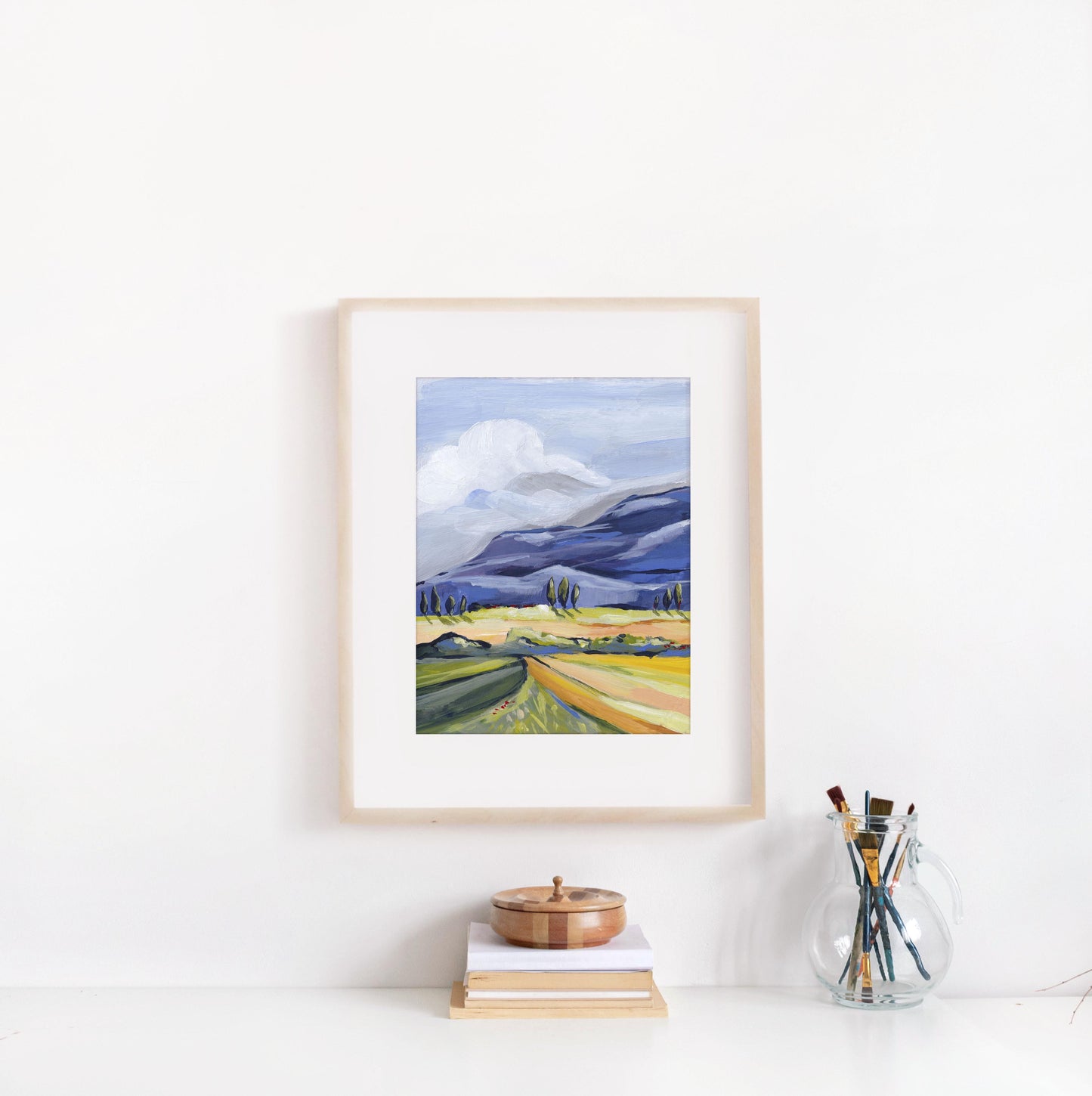 Framed landscape painting on a white wall with books and a glass jar of paintbrushes below.