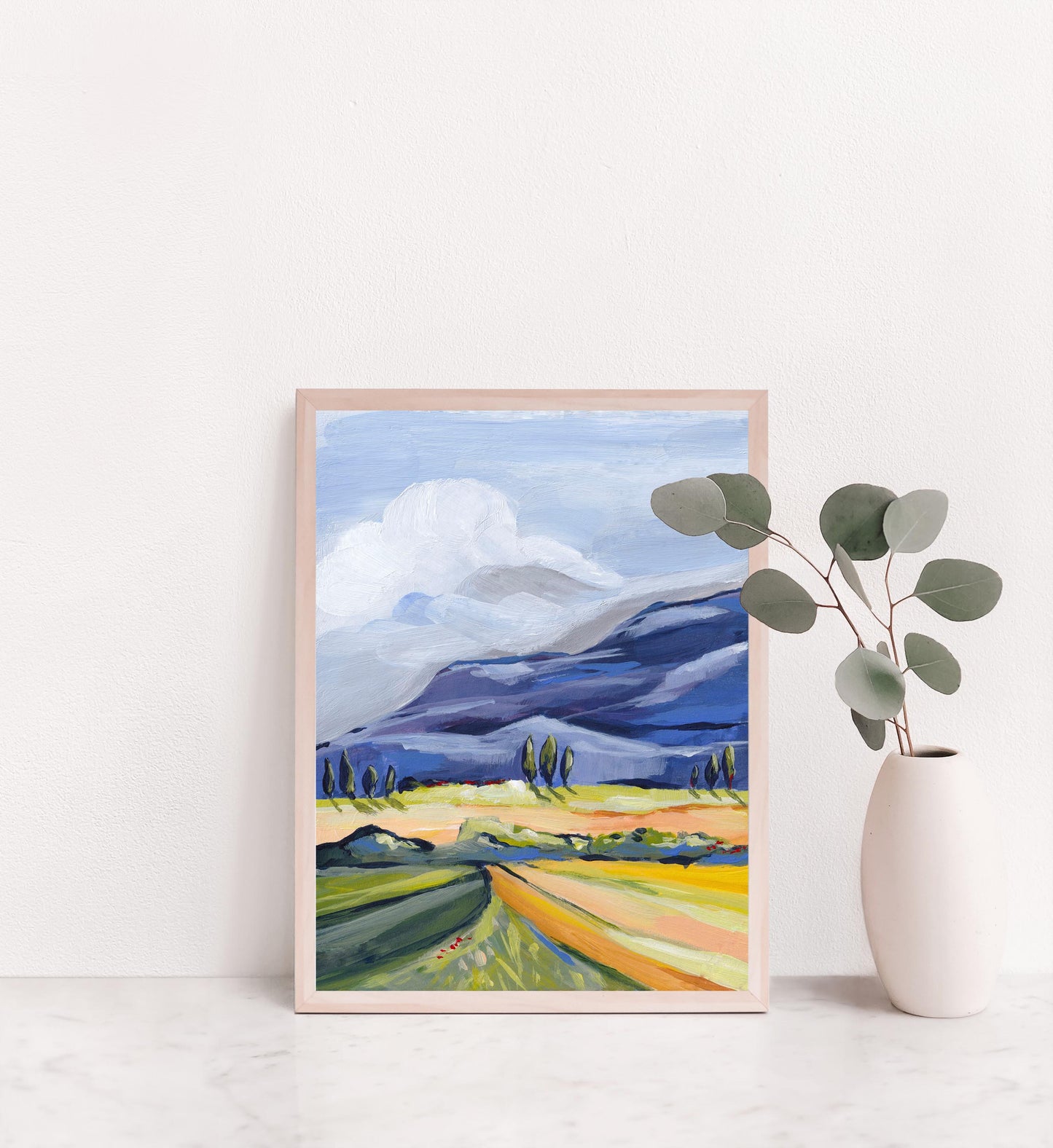 Framed landscape painting with mountains and fields on a white wall.