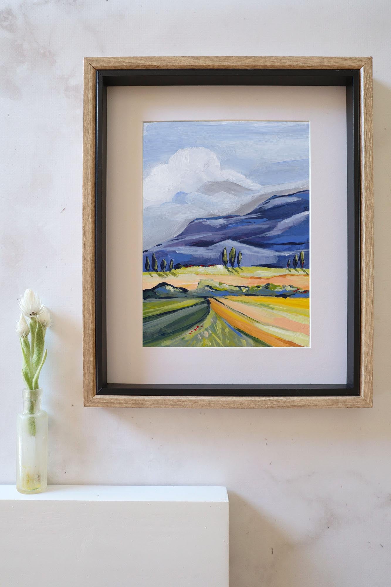 Framed landscape painting on a white wall with a vase of flowers below.