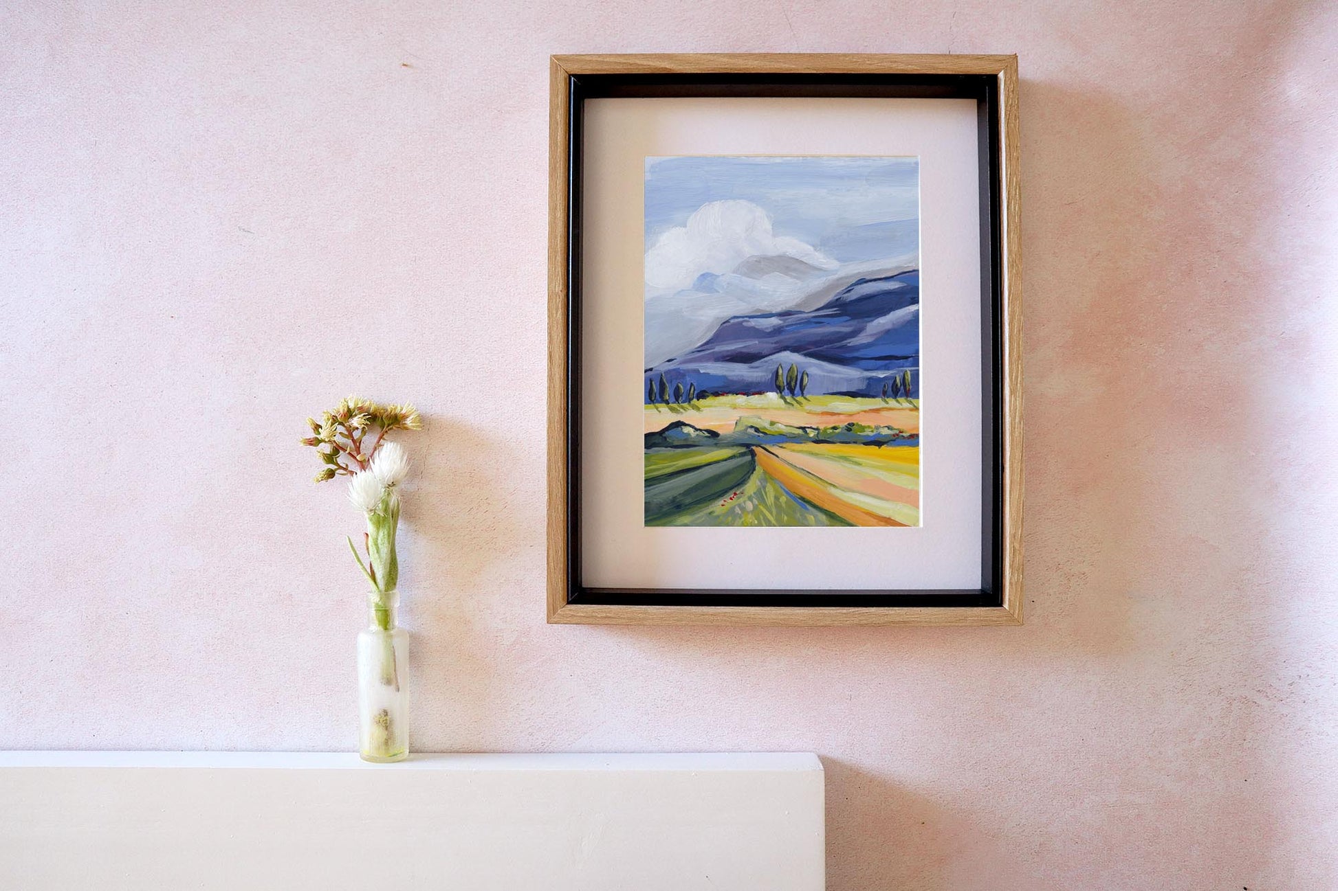 Framed painting of a landscape on a wall with a vase of flowers beside it.