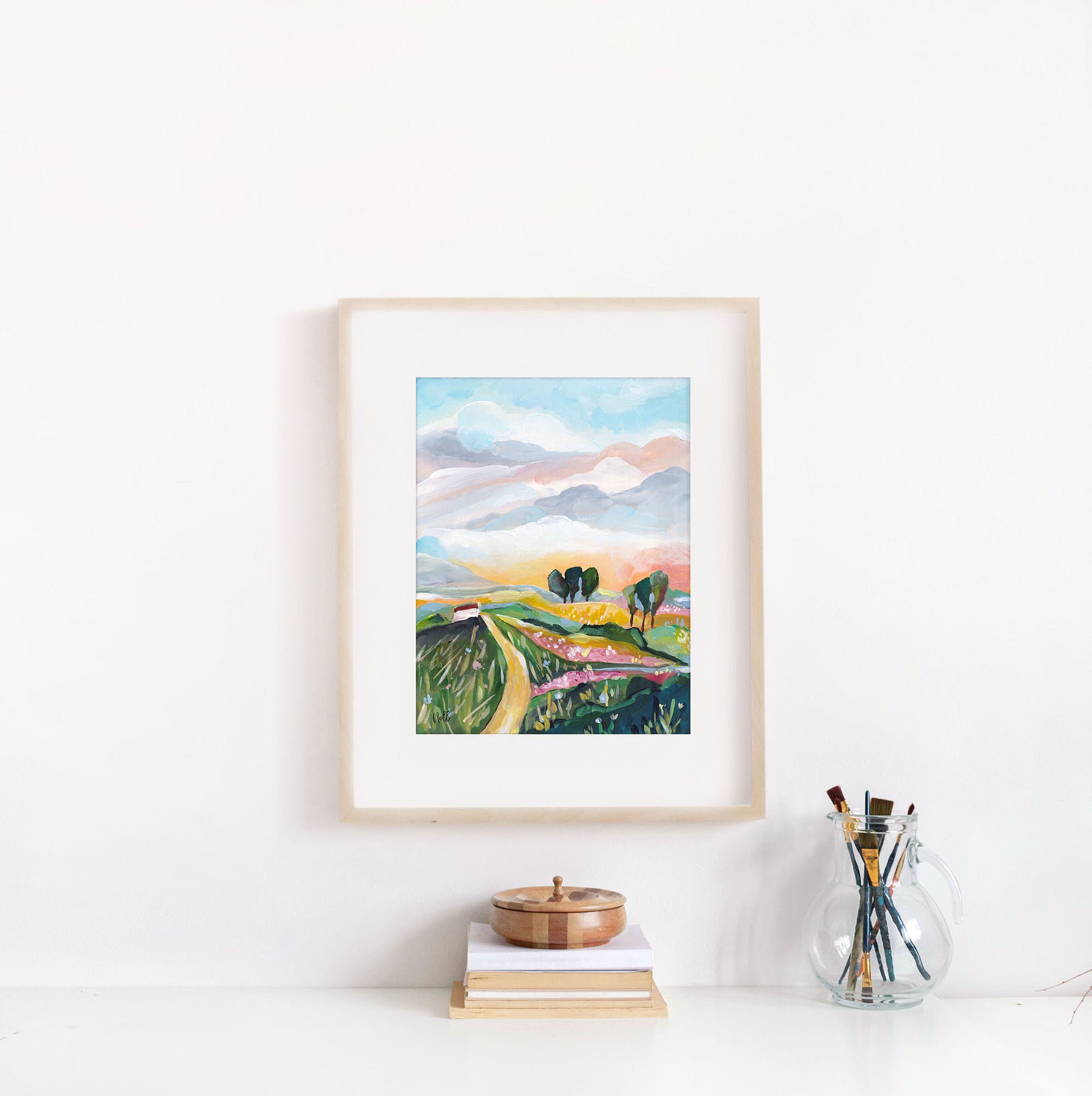 Framed artwork of a landscape with mountains and fields on a white wall.