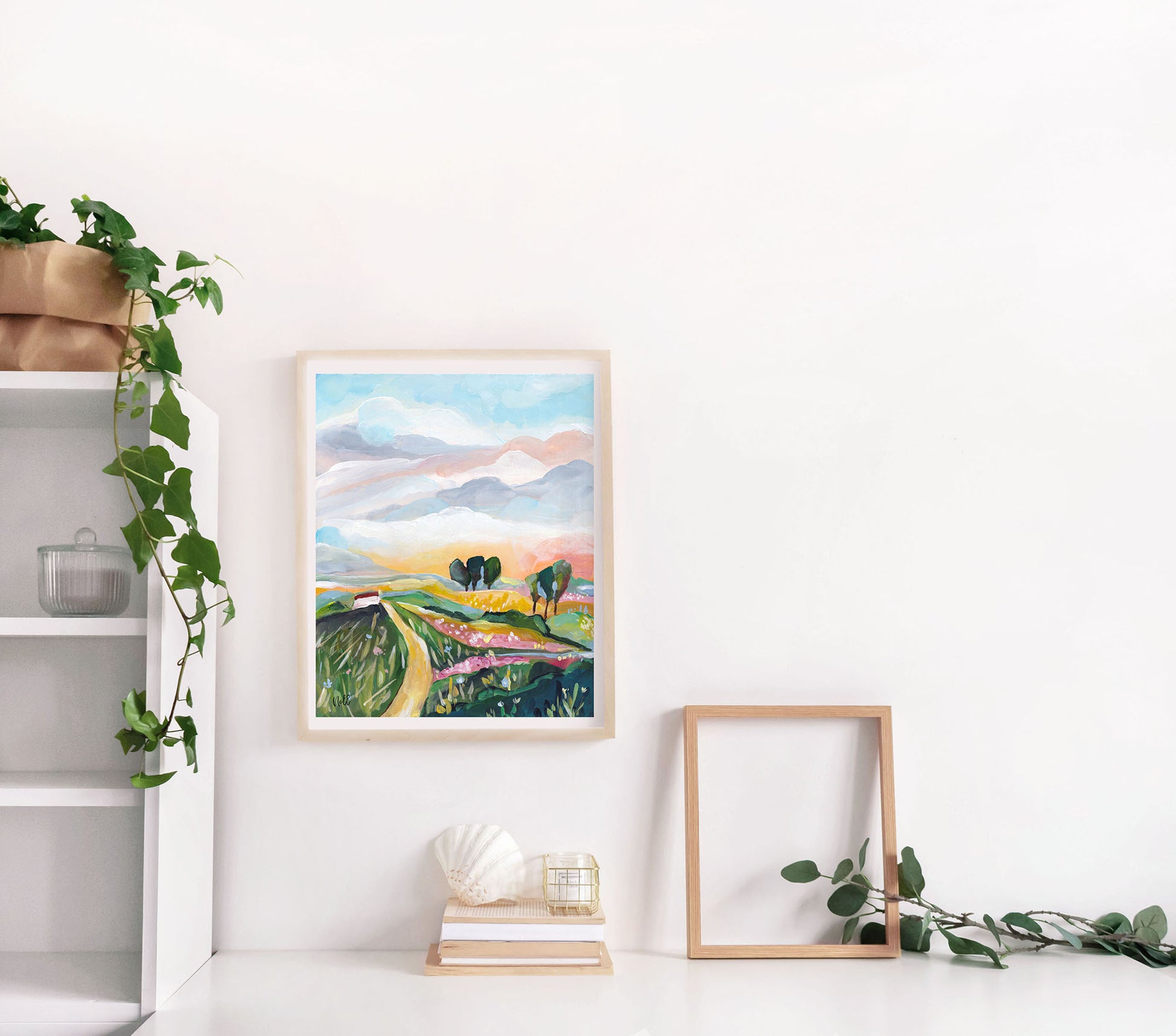 Framed landscape painting on a white wall with a plant and books below.
