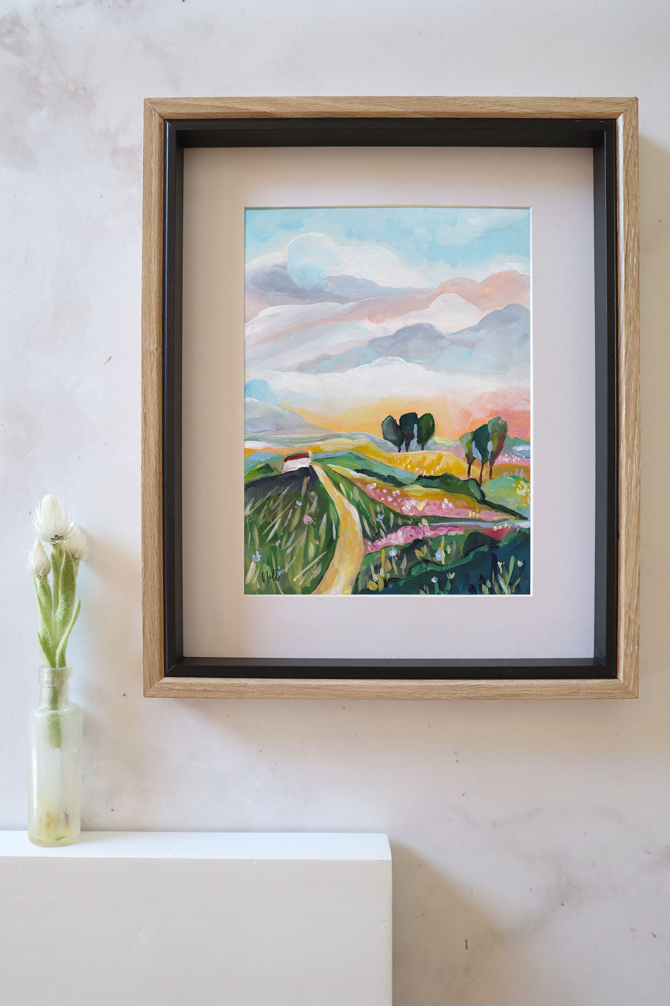 Framed painting of a landscape with mountains and fields on a white wall.